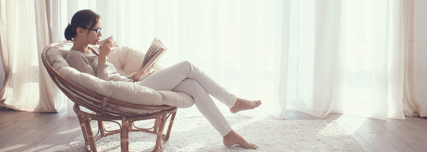 Photo of a woman sitting in a chair, holding a book and drinking a cup of coffee.